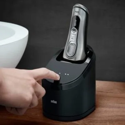 Braun shaver shown charging in its cleaning and charging station, with a finger pressing the button.