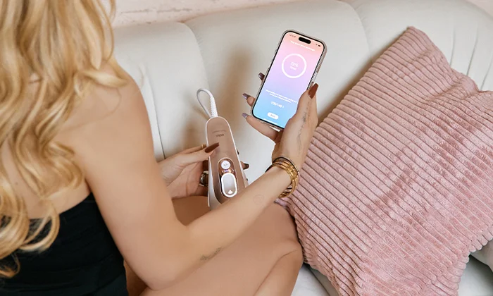 Woman on a couch using an IPL while controlling it with a smartphone app nearby.